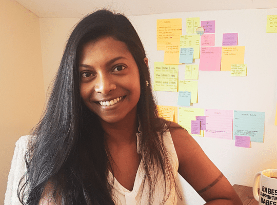 Image of Ruha Thurairatnam, youn woman tech wizard artist entrepreneur of The Designist Studio.  Ruha is smiling and standing in front of a wall filled with sticky notes.