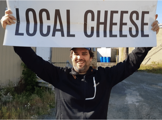 Adam Blanchard of Five Brothers Artisan smiling broadly and holding up a Local Cheese sign