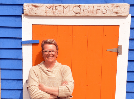 Bizzie Tizzie Book series Author Yvonne Bryant standing in front of a bright fishing stage with orange door in New Perlican NL
