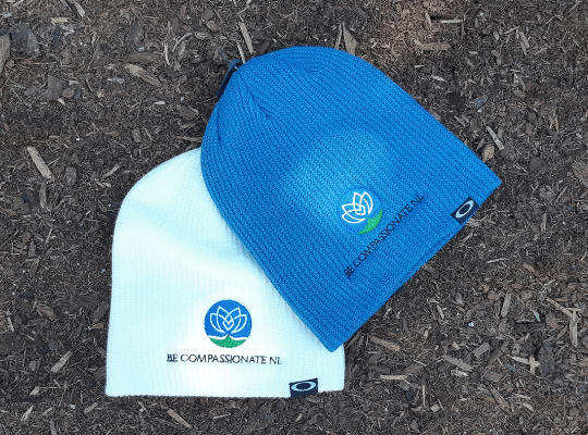 Two beanies one white one blue on the ground with the Be Compassionate NL logo 