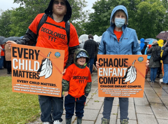 Three people in a row standing outdoors in light rain bearing signs that read Every Child Matters in French and English.  At left, a bearded man in an orange t-shirt with the words Decolonize YYT, centre a smiling child in the same orange t-shirt, right a blonde woman wearing a blue jacket. 