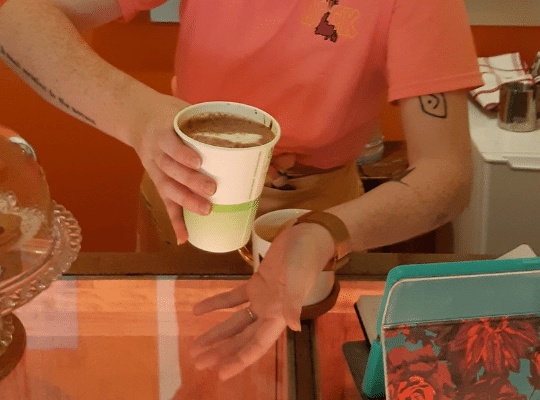 Cappuccino in a compostable cup at Elaine's Cafe.  Two arms are visible and the colours are pinkish/orange.
