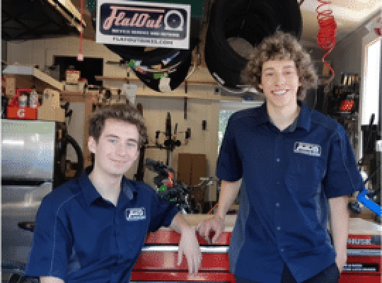 Two young men in blue shirts under the FlatOut sign in the service garage