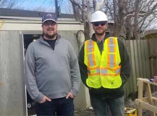 two men, staff at Impact Construction, standing outdoors near a sawhorse. one wearing a hard hat and yellow vest