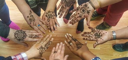 9 women's standing in a circle with on of their hands extended forward to the center and all hand have henna designs on them. some hands are palm up and some are palm down.