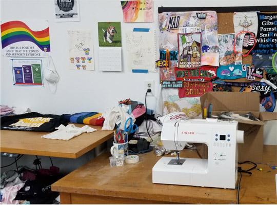 Production at The Shop:  a sewing machine on a desk in front of a wall full of pictures and a pride flag