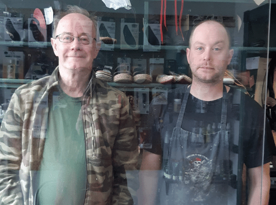 Two men standing behind plexiglass counter ready to help at Modern Shoe Hospital 