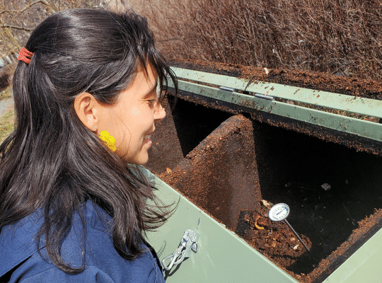 Smiling dark-haired woman standing outdoors in front of a compost bin. Vivana Rameriz Luna, Founder of Planeet.