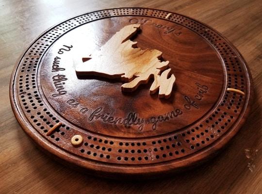 round dark wooden crib board designed with a carving of the island of Newfoundland in the middle and the words 'no such thing as a friendly game of crib'