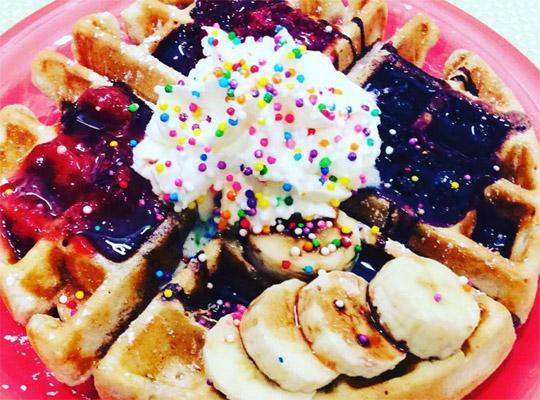 a waffle with berry toppings, sliced banana, chocolate sauce, whipped cream, and sprinkles