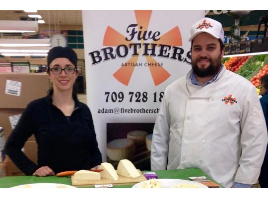 two cheese makers with their cheese on display standing in front of a five brothers poster