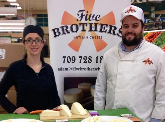 two cheese makers with their cheese on display standing in front of a five brothers poster