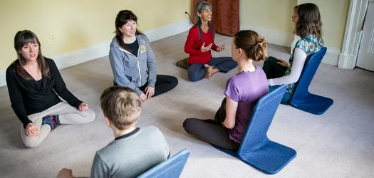 there are 2 rows of people faceing each other on facing and 3 back on in the picture. the ones sitting back on have upright seats to help positioning of their backs, they are all sitting with their feet tucked underneath them in a yoga position and they all look like they are engaged in conversation. They are all sitting on gray carpet in a room that is painted yellow