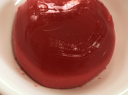 A round scoop of my mother's three-layered jello that I helpped her make. The jello is cherry coloured and favoured. It is sitting in a white bowl.