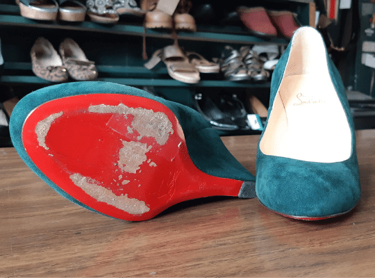 a pair of teal suede Christian Louboutin shoes rest on a counter awaiting repair of their signature red soles