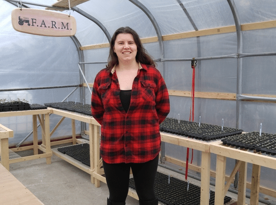 smiling dark-haired woman in a red shirt in a greenhouse surrounded by flats of seedlings 
