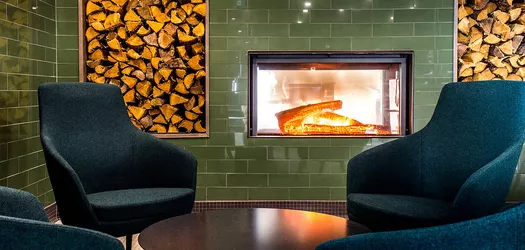4 material covered chairs around a round dark wooden table and in the back ground in a green tiled wall with a lite fireplace in the middle of it and there are 2 stock piles of wood, one on each side of the fire place. 